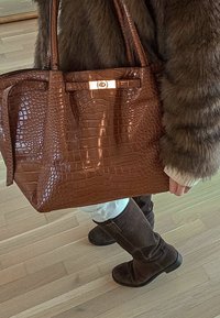 Personne portant des bottes marron foncé et un manteau en fourrure, tenant un grand sac à main en cuir brun texturé avec des fermetures dorées sur un sol en bois.