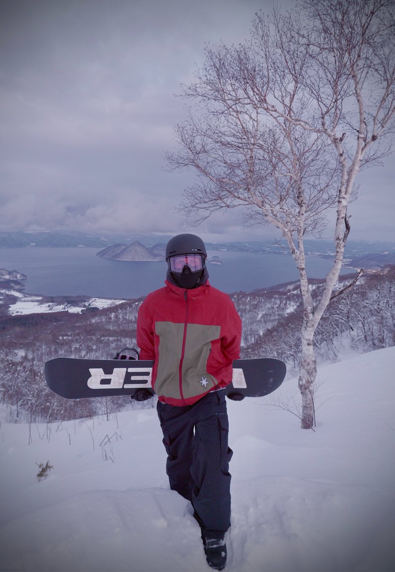 Personne portant une veste rouge et grise, un casque noir et des lunettes de protection, tenant un snowboard, debout sur une pente enneigée avec un arbre dépouillé et un lac en arrière-plan.
