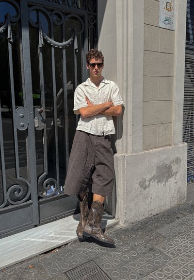 Botas de cowboy de cuero marrón, camiseta blanca de manga corta y ligera, y pantalones cortos oscuros y holgados, apoyándose contra una puerta de metal decorativa.