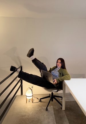 Joven sentada en una silla de oficina con un portátil, con las piernas levantadas y apoyadas en la barandilla, junto a una mesa blanca y una lámpara de pie.