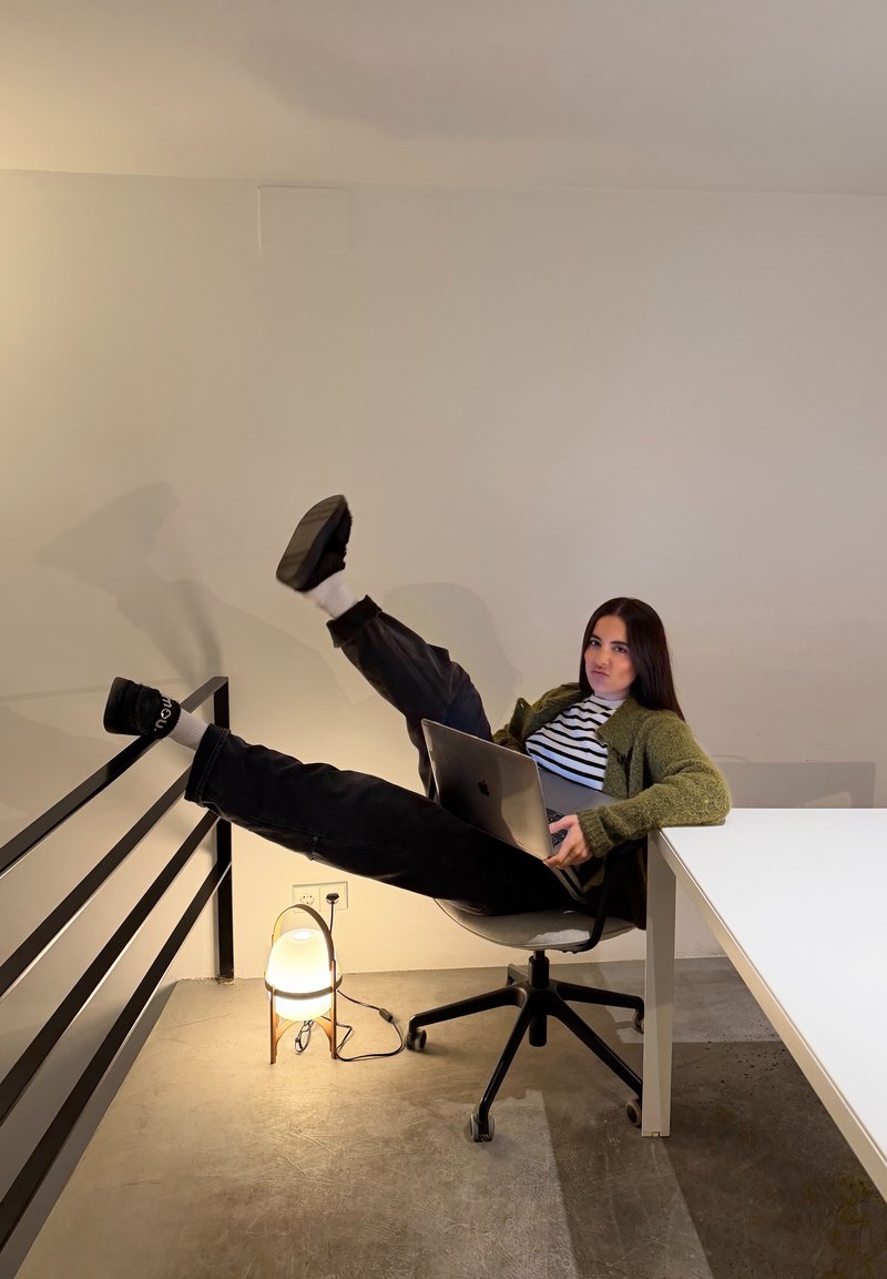 Jeune femme assise sur une chaise de bureau avec un ordinateur portable, jambes levées et reposant sur une rampe, à côté d'une table blanche et d'une lampe sur pied.