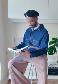 Homem a usar um boné preto e uma jaqueta de ganga sentado num banco a ler um livro numa sala iluminada com uma planta e um alto-falante nas proximidades.