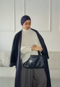 Femme portant un foulard bleu marine et un manteau avec un pull crème et un pantalon gris, tenant un sac à main noir, debout dans une pièce blanche minimaliste.