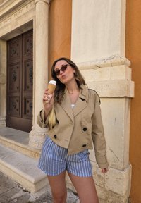 Un abrigo trench beige claro con botones negros, pantalones cortos a rayas azules y blancas, sosteniendo un cono de helado, de pie cerca de una pared naranja texturizada.