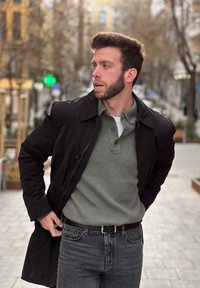 Cappotto nero con bottoni sopra un polo a coste verde chiaro. Jeans scuri con una cintura nera. Pavimentazione liscia e sfondo sfocato di una strada.