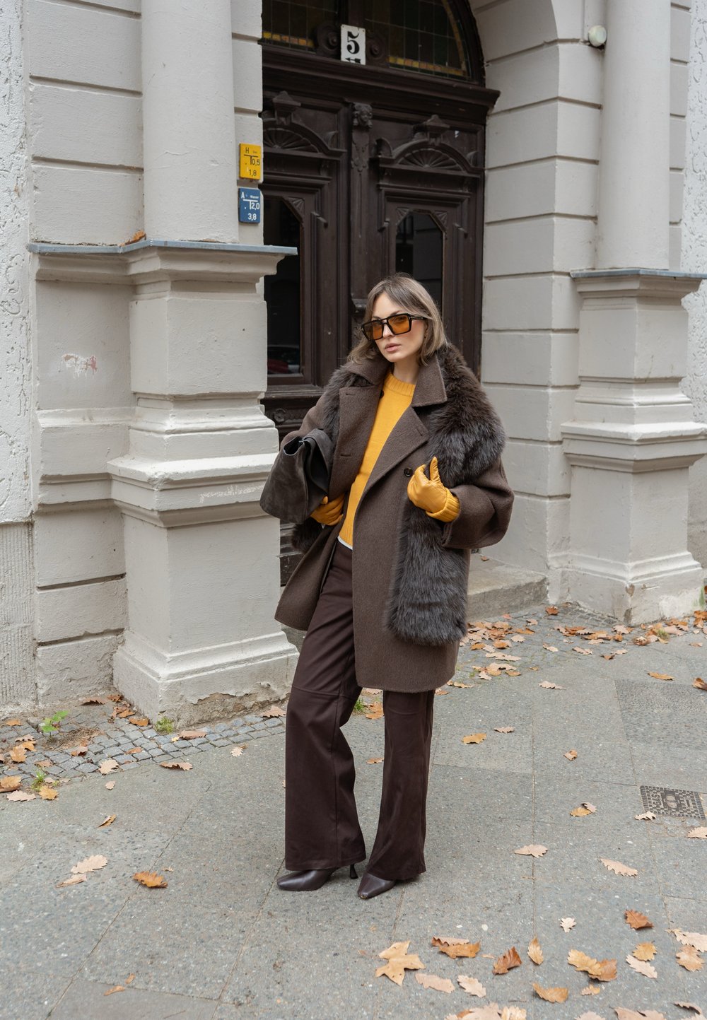 Barna gyapjúkabát műszőrmés gallérral, mustársárga pulóver, barna bőszárú nadrág és sárga kesztyűk. Csillogó napszemüveg egészíti ki a szettet.