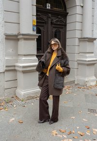 Barna gyapjúkabát műszőrmés gallérral, mustársárga pulóver, barna bőszárú nadrág és sárga kesztyűk. Csillogó napszemüveg egészíti ki a szettet.