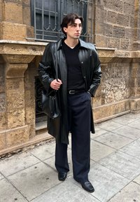 Jeune homme portant un manteau en cuir texturé noir, un pull noir, un pantalon large à fines rayures bleu marine, des chaussures noires et portant un sac en cuir noir dans une rue pavée.