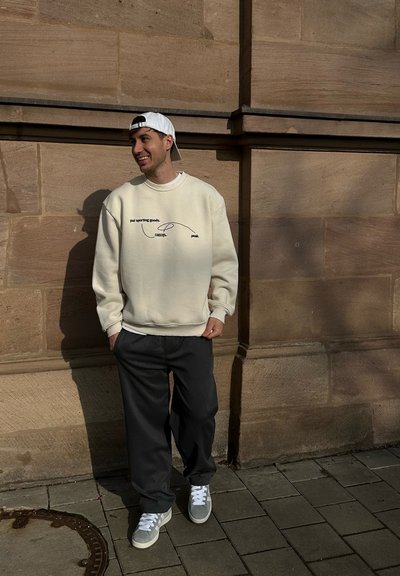 Sudadera crema con texto y gráfico en negro, combinada con pantalones oscuros y zapatillas grises. El fondo presenta paredes de piedra y pavimento.