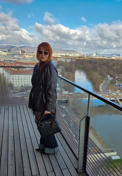 Mujer con cabello rojo que lleva una chaqueta de cuero negra y gafas de sol, de pie en un balcón con vistas a la ciudad, el río y un cielo nublado.