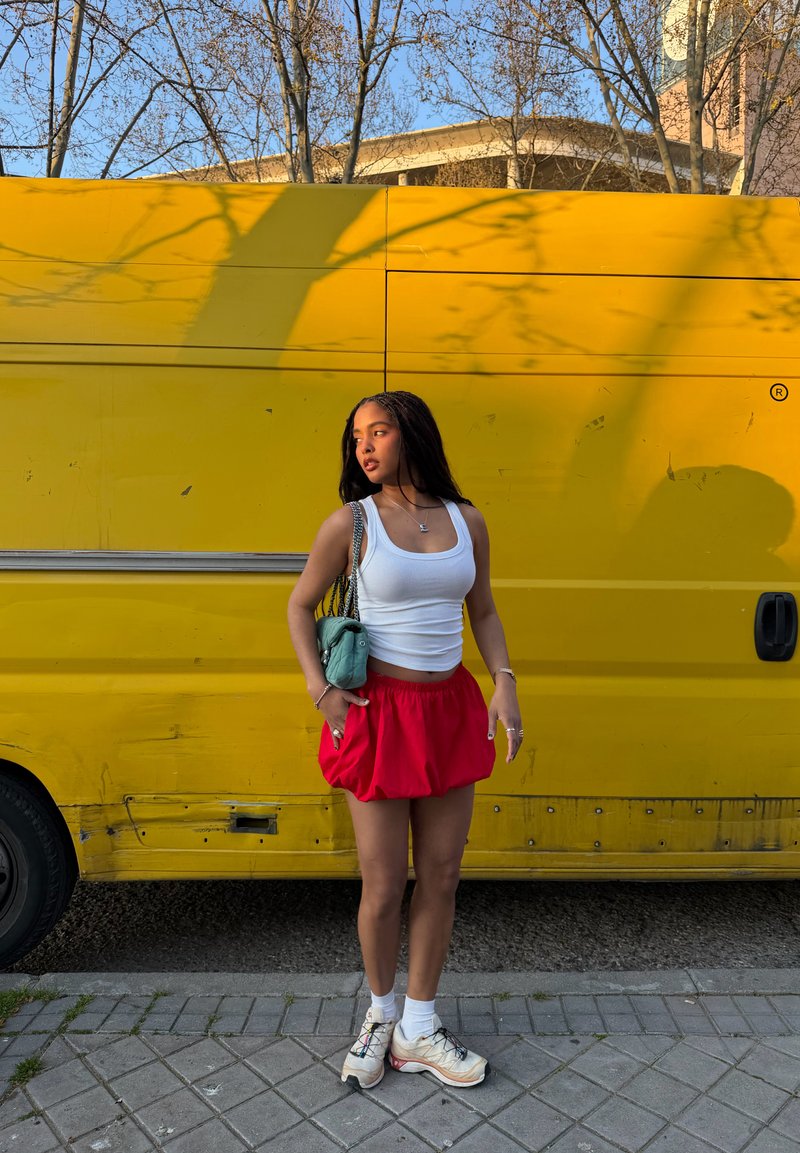Young woman in white tank top, red skirt, white sneakers, and turquoise bag stands on sidewalk in front of yellow van under leafless trees.