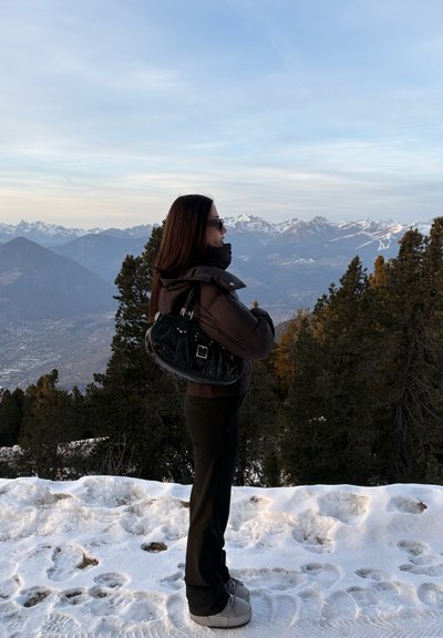 Mujer con gafas de sol y chaqueta de invierno de pie sobre suelo nevado, mirando hacia una cadena montañosa con pinos al atardecer.