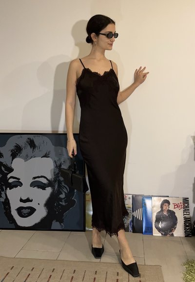 Mujer con vestido de tirantes negro y gafas de sol posa en el interior cerca de discos de vinilo y una lámina enmarcada de arte pop de Marilyn Monroe.