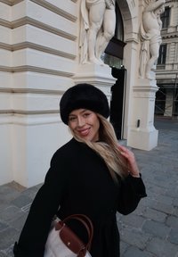 Černá kožichová čepice nošená s černým kabátem, béžová taška s hnědými akcenty. V pozadí je zdobená bílá architektura a kamenná dlažba.