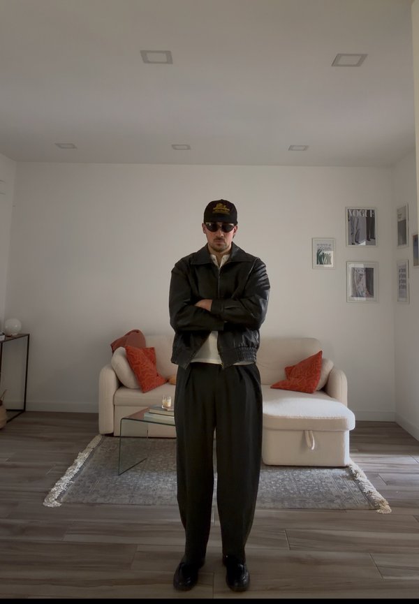 Mann iført svart skinnjakke, caps og solbriller står med armene krysset i en stue med beige sofa og røde puter.