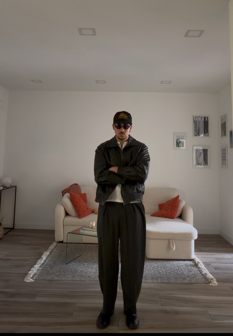 Mand iført sort læderjakke, kasket og solbriller står med armene over kors i en stue med beige sofa og røde puder.