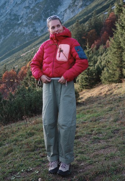 Mujer con chaqueta de plumas roja, botas de senderismo grises y pantalones holgados se encuentra en una ladera cubierta de hierba con montañas boscosas de fondo.