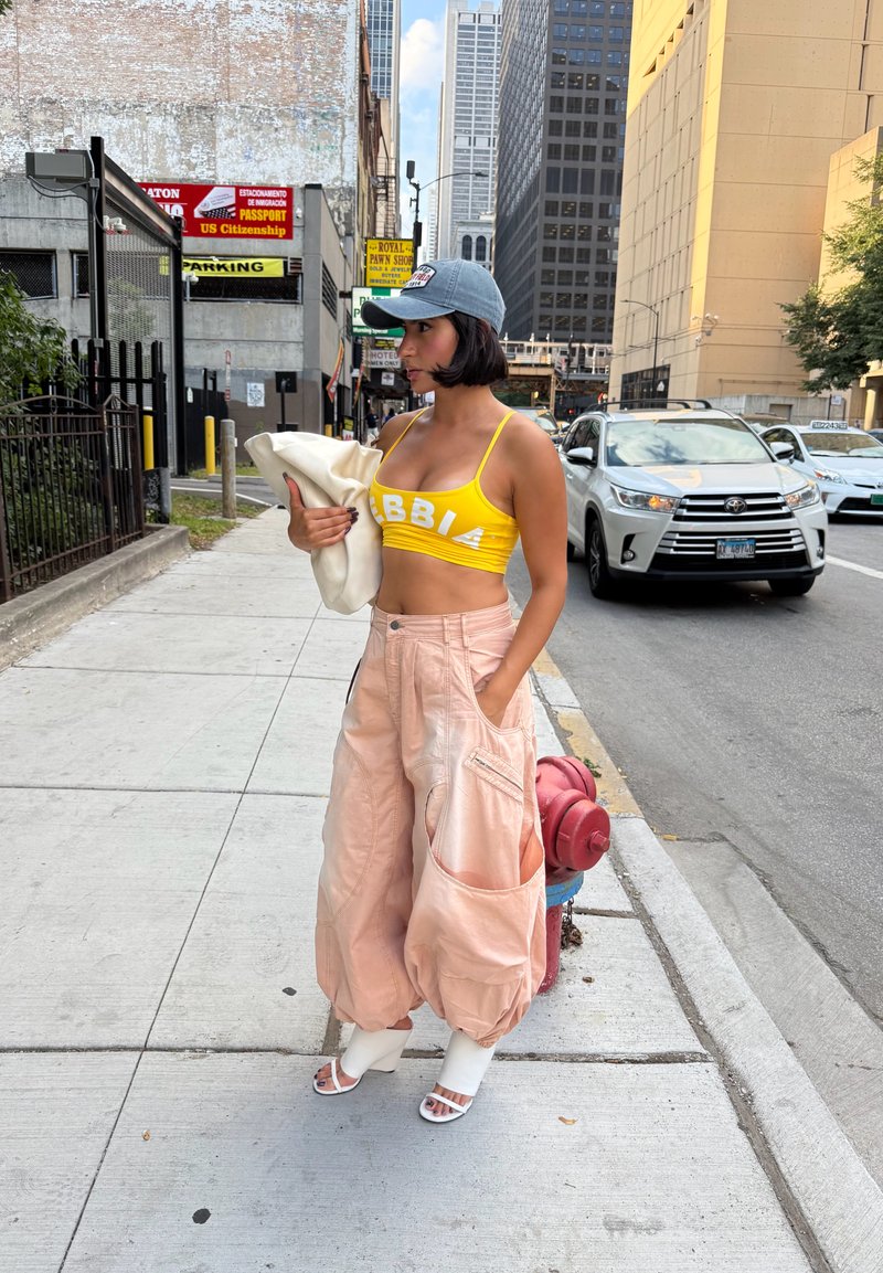 Mulher com top amarelo e calças largas rosas segurando uma jaqueta branca, em pé na calçada da cidade, perto de um hidrante vermelho e de um SUV branco.