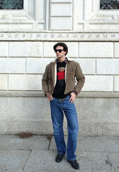 Chaqueta de ante marrón con cuello de borreguillo, suéter negro con un diseño de oso, vaqueros azules y zapatos negros, contra una pared blanca texturizada.