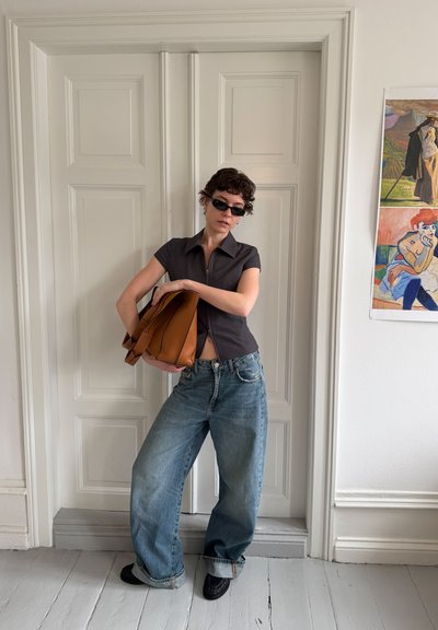 Persona con gafas de sol, camiseta negra de manga corta y vaqueros anchos azules, sosteniendo una gran bolsa marrón, parada frente a unas puertas dobles blancas.