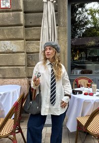 Hvit oversized skjorte, marineblå stripete slips, grå beret, og mørke denim bukser. Holder et kamera; svart veske over skulderen, kafé scene i bakgrunnen.