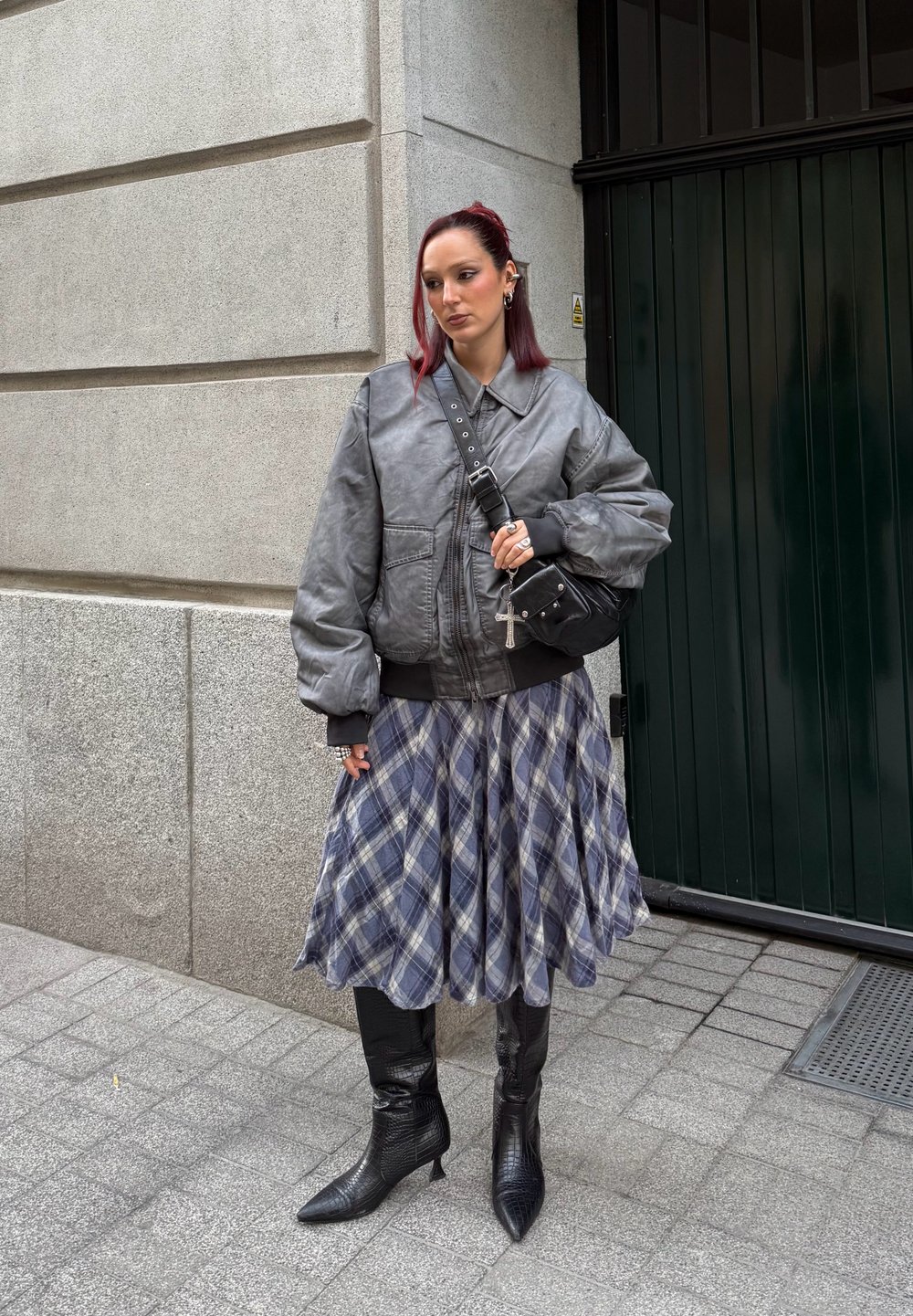 Veste bomber grise, jupe à carreaux bleue et blanche, bottes noires montantes avec des bouts pointus, et sac bandoulière avec des accents cloutés.