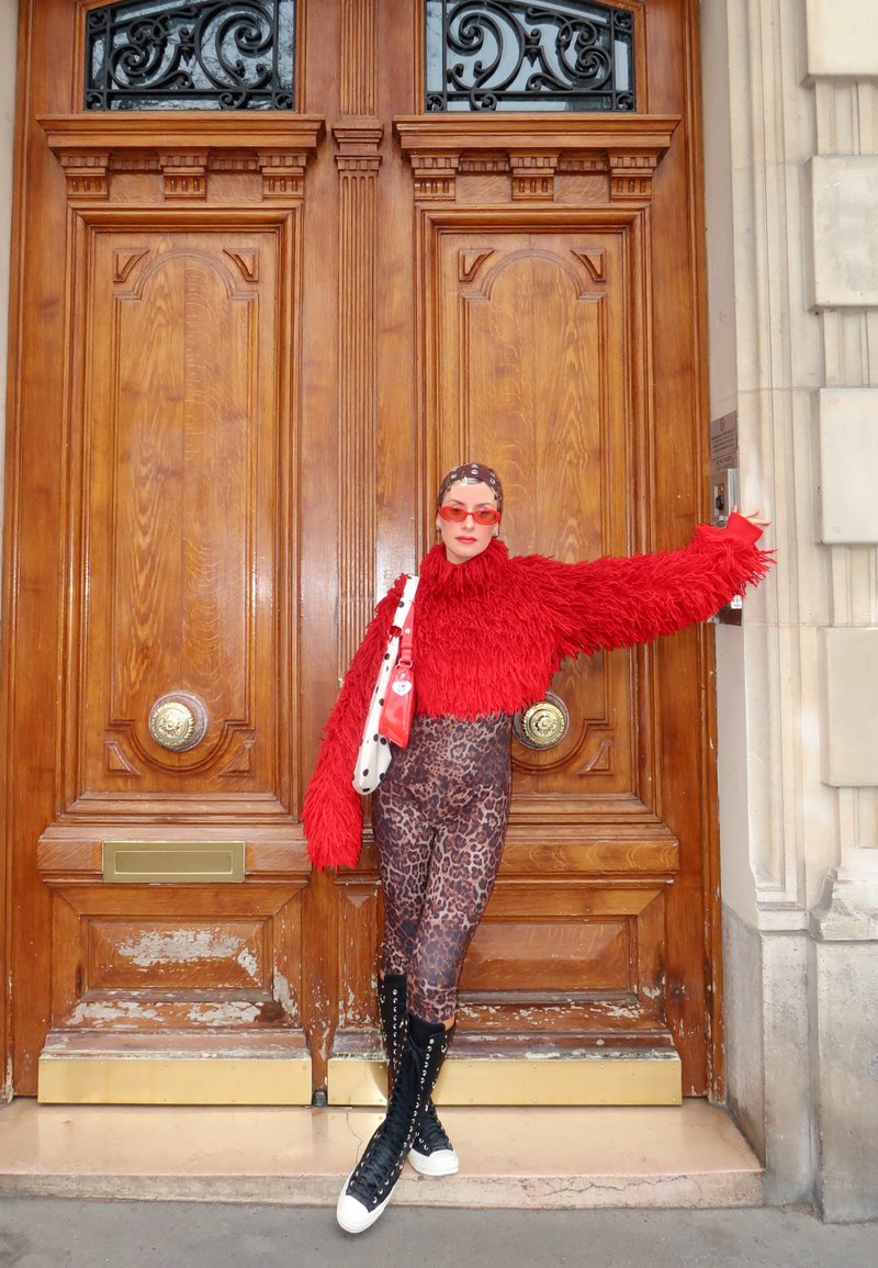 Person in red fleecy jumper, leopard-print leggings, and black high-top trainers leaning against large ornate wooden doors.