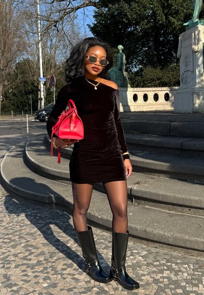 Vestido negro de terciopelo sin hombros, bolso rojo con herrajes dorados, botas negras hasta la rodilla y gafas de sol redondas, todo contra un fondo de piedra.