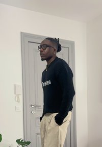 Homme portant des lunettes et des boucles d'oreilles, vêtu d'un pull noir avec le texte "Timeless" et d'un pantalon beige, debout à l'intérieur près d'une porte grise, les mains dans les poches.