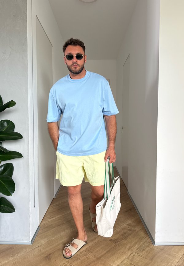 Hombre con gafas de sol, camisa azul clara, pantalones cortos amarillos, sandalias beige, llevando una bolsa blanca caminando en el interior sobre un suelo de madera.