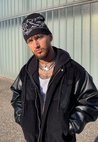 Homme portant un bonnet noir et une veste noire avec des manches en cuir, debout à l'extérieur contre un mur en verre givré, sous la lumière du soleil.