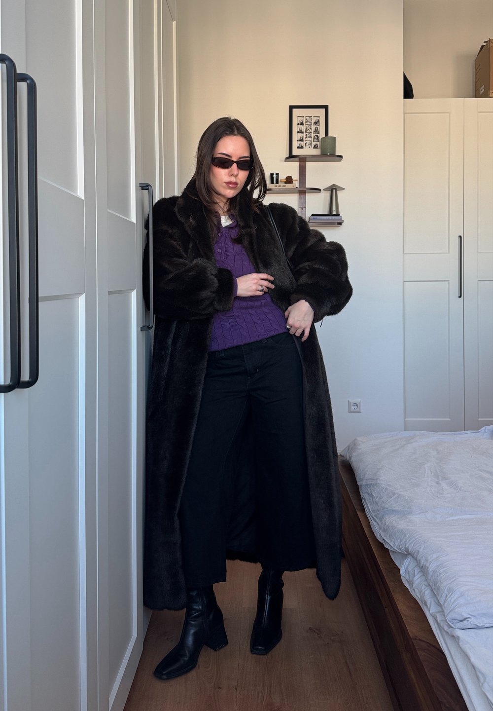 Long manteau en fausse fourrure noire porté sur un pull en tricot violet, pantalon noir à jambes larges et bottines noires à talons. Accessoires incluant de petites lunettes de soleil.