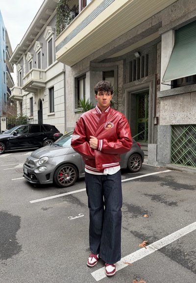 Joven de pie en un estacionamiento de la ciudad cerca de coches, vistiendo una chaqueta varsity roja, vaqueros oscuros y zapatillas rojas con blanco.