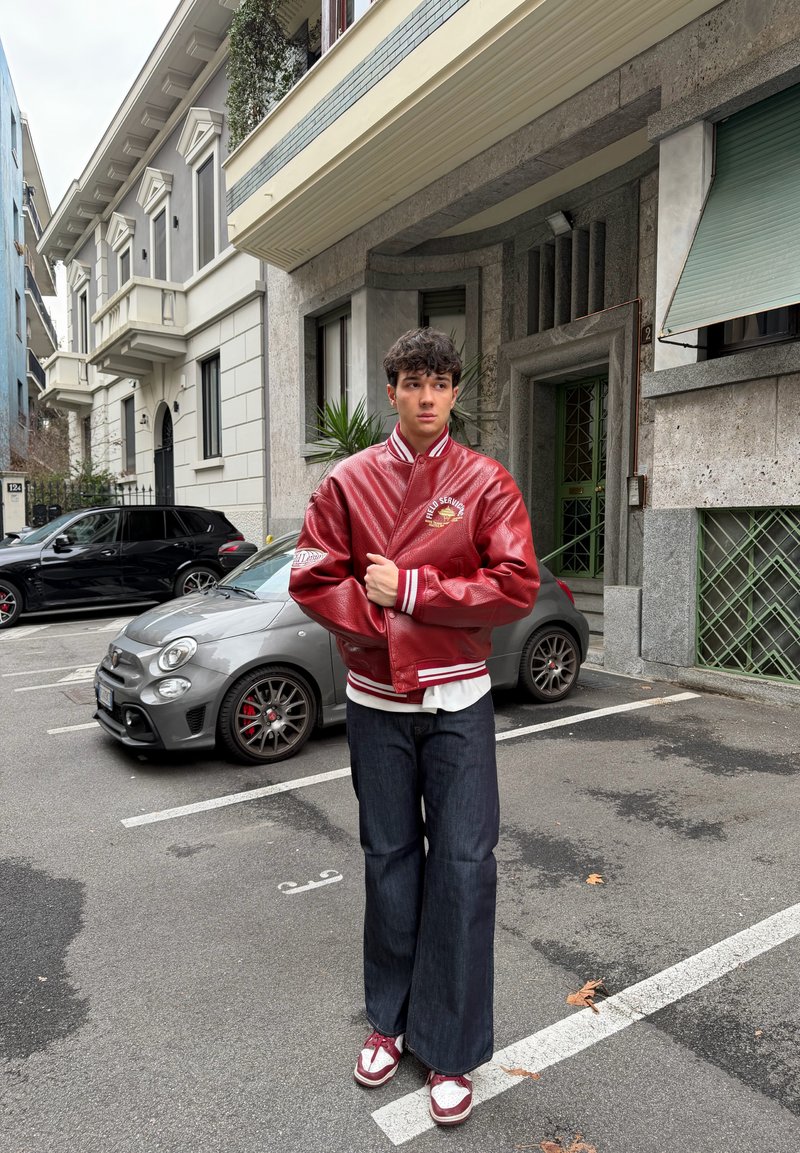 Jeune homme debout dans un parking en ville près de voitures, portant une veste universitaire rouge, un jean foncé et des baskets rouges et blanches.