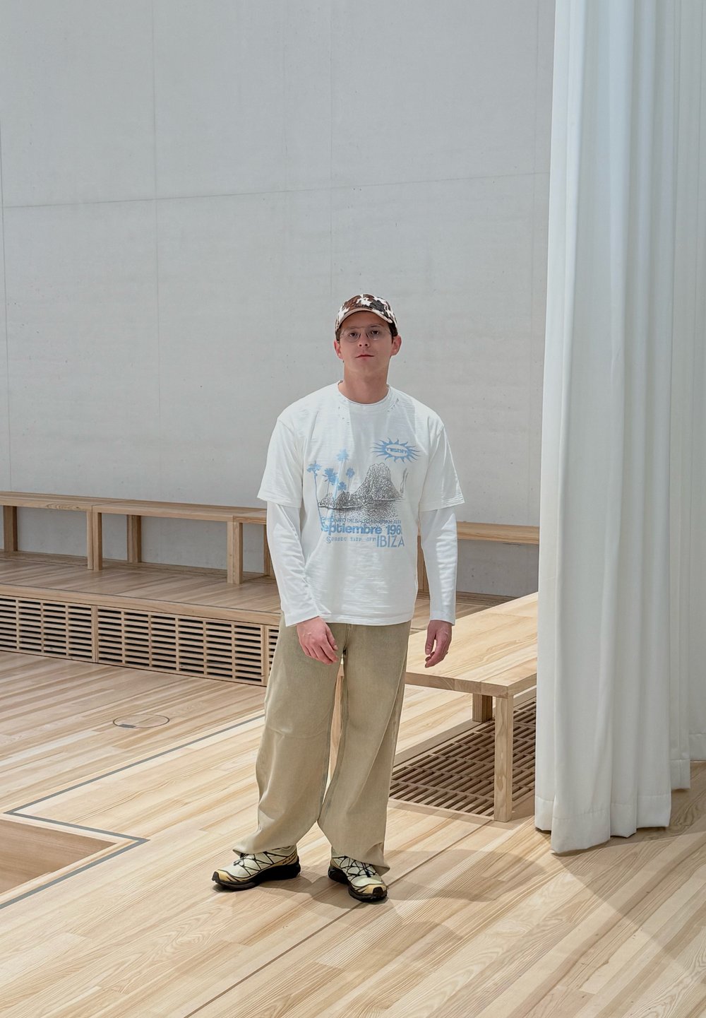 Un jeune homme portant des lunettes, une casquette camouflage, un t-shirt blanc à manches longues avec un motif graphique, un pantalon beige et des baskets, debout dans une pièce en bois minimaliste.