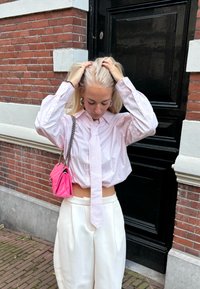 Ružovobiele pruhované krátke tričko s kravatou, v kombinácii s bielymi voľnými nohavicami. Drží ružovú prešívanú kabelku s reťazovým remienkom.