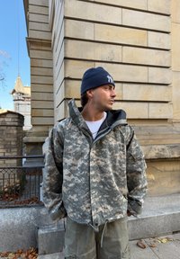 Pelēka krekliņa jaka ar augstu apkakli, izteikti teksturēta. Valkāta virs baltas krekli�ņa, kombinēta ar gaiši zaļām kaujas biksēm.