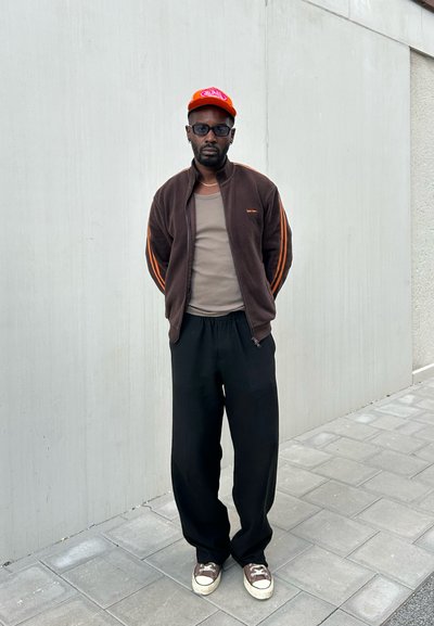 Chaqueta de forro polar marrón con rayas naranjas, camiseta sin mangas marrón claro, pantalones negros de corte holgado y zapatillas marrones con suelas blancas, complementados con una gorra naranja.