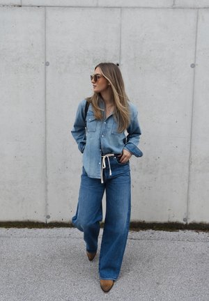 Camisa de denim y jeans de pierna ancha en diferentes tonos de azul, complementados por zapatos marrones. La camisa presenta bolsillos y un diseño de botones.