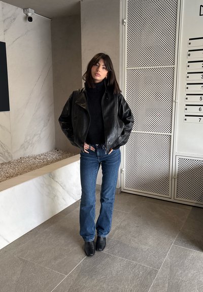 Chaqueta corta de cuero negro sobre un suéter oscuro de punto, combinada con vaqueros azules acampanados y botines negros. Fondo de un interior moderno.