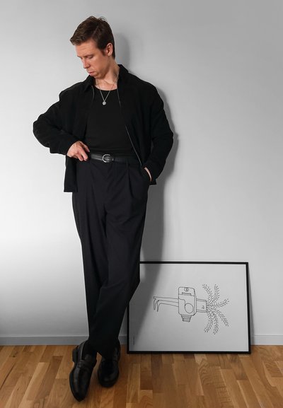 Chaqueta negra sobre una camisa negra, pantalones oscuros y zapatos negros. Ajuste slim, textura suave, de pie junto a una obra de arte minimalista enmarcada.