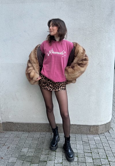 Camiseta de algodón rosa con el texto "Dramático", falda corta de leopardo, medias negras transparentes y botas negras con cordones, todo superpuesto con un abrigo de piel marrón.