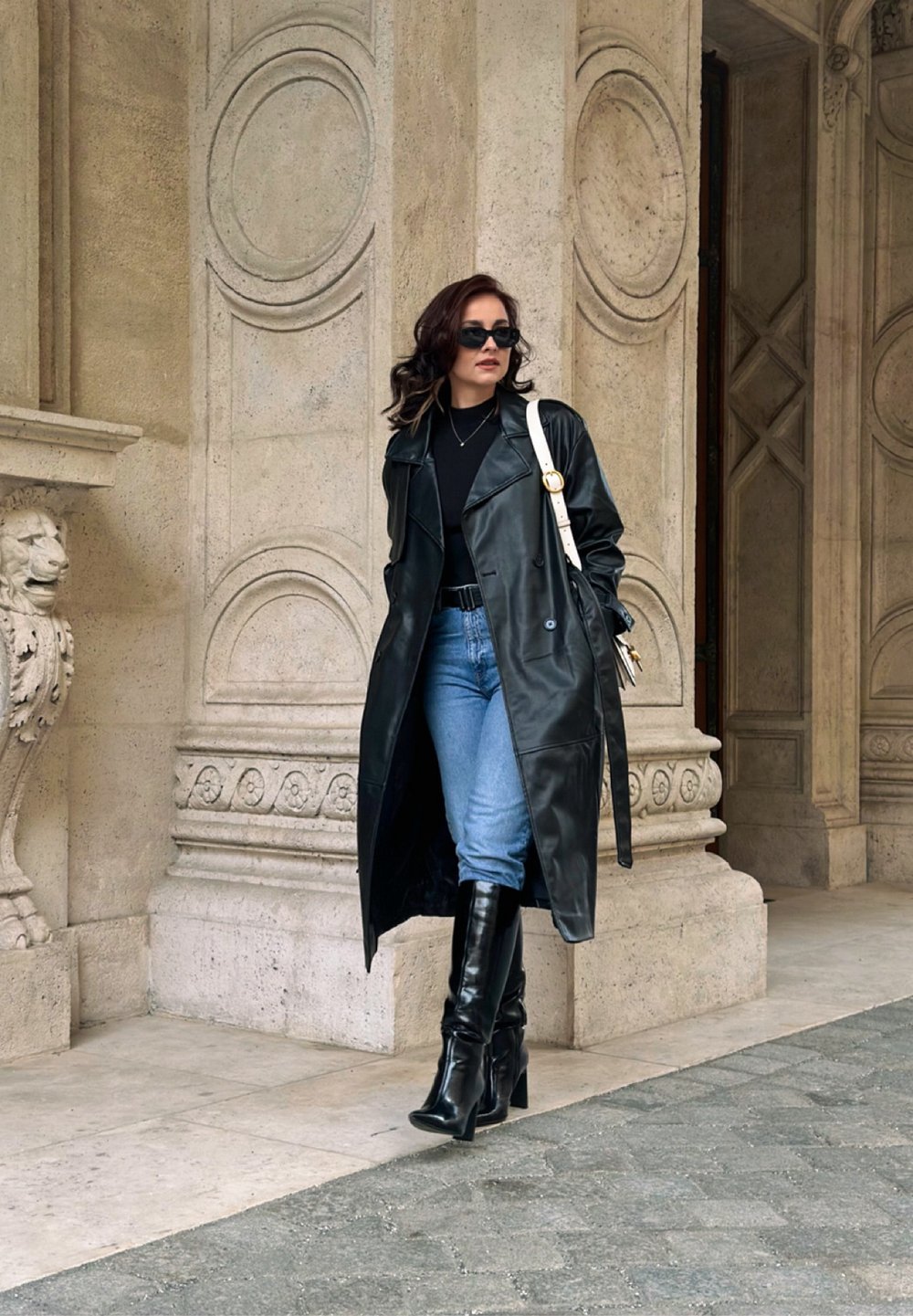 Cappotto trench in pelle nera, top nero attillato, jeans azzurri chiari e stivali neri con tacco al ginocchio. Borsa bianca con un accento dorato.