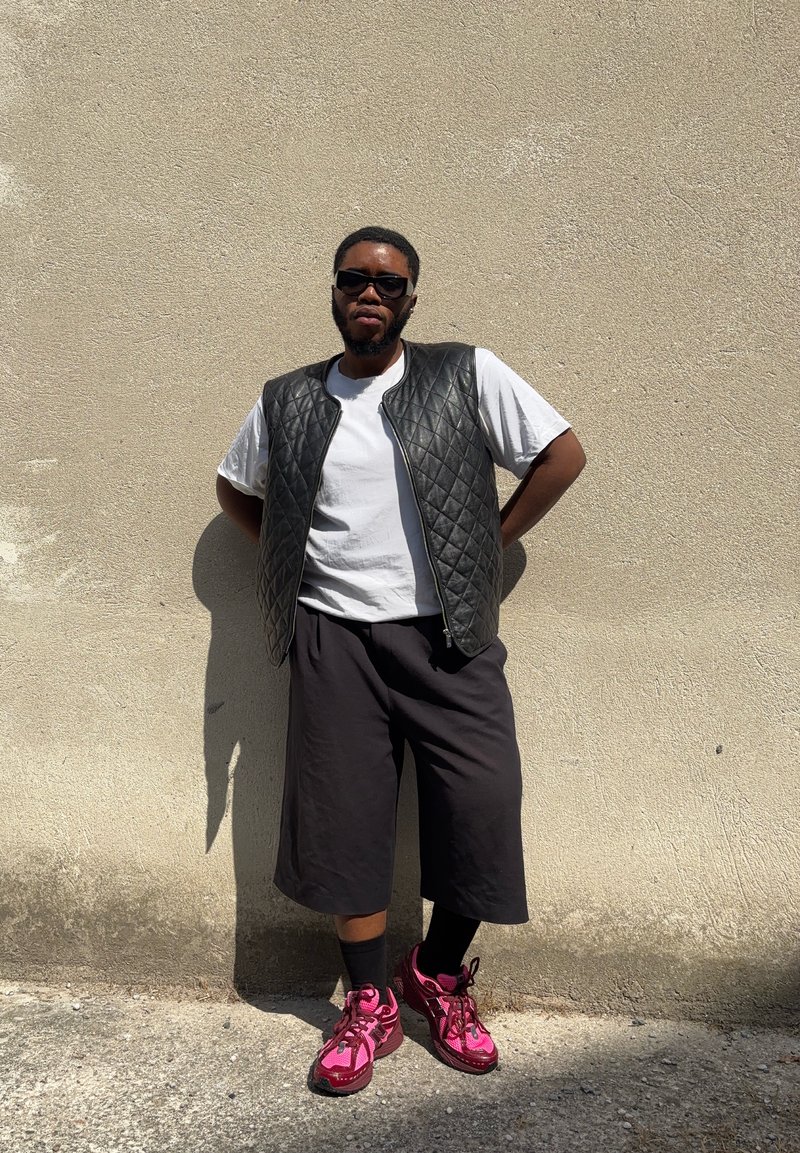Gilet trapuntato nero, t-shirt bianca, pantaloni corti neri larghi e sneakers rosa. In piedi contro un muro chiaro con texture.