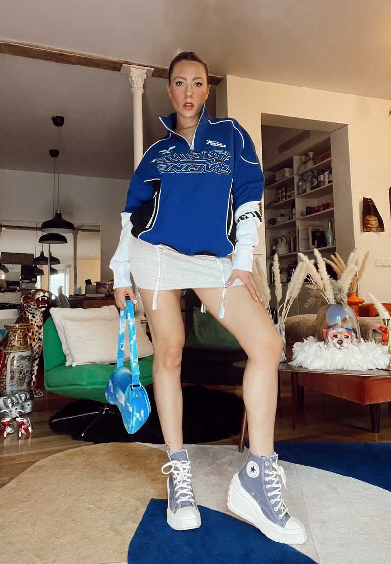 Pullover azul com detalhes brancos, saia mini cinza com cordões, mala azul e ténis altos cinzentos com solas brancas.