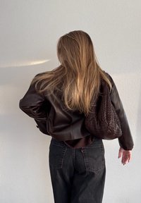 Femme aux longs cheveux blonds portant une veste en cuir sombre et un jean noir, avec un sac à main tissé foncé sur l'épaule, face à un mur clair.