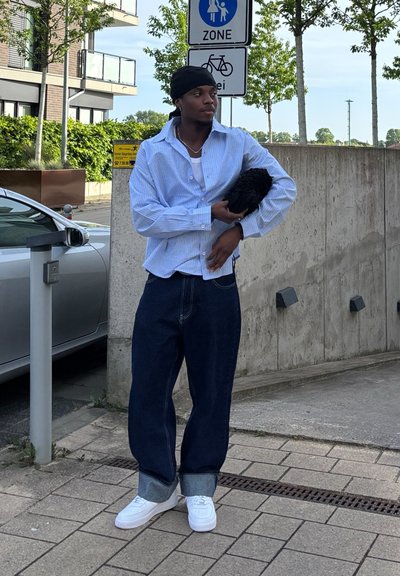 Camisa de botones a rayas azules, jeans anchos oscuros con dobladillo, zapatillas blancas, sosteniendo un objeto negro y peludo. Entorno urbano con letreros y vegetación.