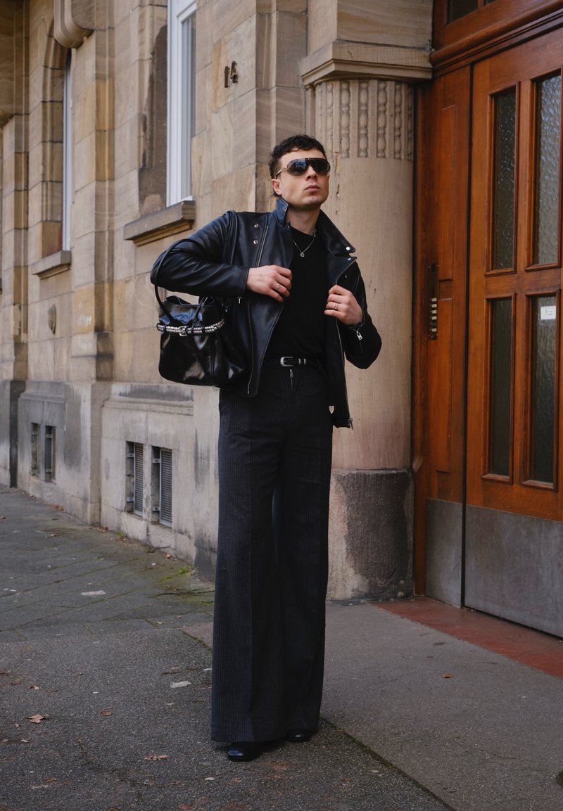 Mann trägt schwarze Lederjacke, Sonnenbrille, weiter gestreifte Hose und trägt eine schwarze Handtasche, steht in der Nähe des Eingangs eines Steingebäudes.