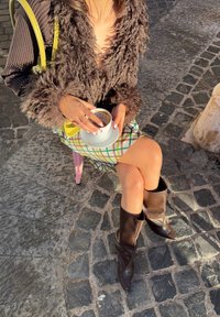 Brauner flauschiger Blazer mit strukturierten Ärmeln, karierter Minirock in Grün- und Gelbtönen und kniehohe braune spitze Stiefel. Hält eine weiße Tasse.