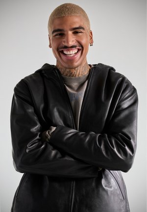 Chaqueta de cuero negra con capucha, textura suave y cremallera frontal. Usada sobre una sudadera gris con bordado azul. Sujeto masculino sonriendo.